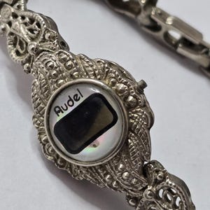 May include: Close-up of a silver-toned bracelet with ornate, detailed links. The central element features a circular design with the word "Audel" and a black rectangle. The bracelet has a decorative, vintage aesthetic.