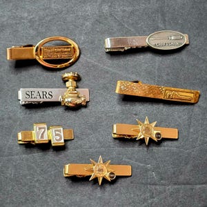 May include: A collection of seven metal tie bars featuring various designs including a gold oval with text, a silver bar with a screwdriver icon labeled Craftsman, a silver bar with a gold faucet and Sears text, a textured gold bar, and star emblems.
