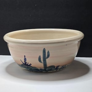May include: A round ceramic bowl with a cream-coloured interior and a smooth rim. The exterior features a soft gradient of peach and cream tones, decorated with hand-painted blue desert imagery including a central saguaro cactus and rocks.