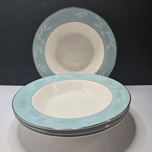 May include: A set of three round ceramic soup bowls featuring a wide, light blue rim decorated with a white, leafy botanical pattern. The centre of each bowl is a solid off-white colour, and the outer edge of the rim is finished with a thin silver band.