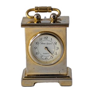 May include: A small, rectangular quartz clock with a gold-colored frame and handle. The clock face is white with black numerals and the words "Times Square" printed on it. The clock is labeled "Japan Movt".