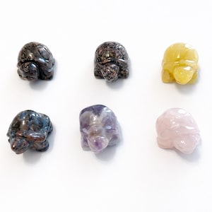 Puede incluir: Seis pequeñas figuras de tortugas de piedra tallada en varios colores. Las tortugas son en tonos de gris, amarillo, morado y rosa. Las figuras se exhiben sobre un fondo blanco.