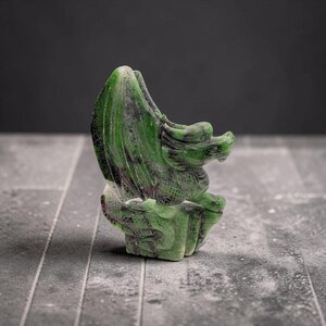以下が含まれることがあります： 目立つ翼と質感のある体を持つドラゴンをかたどった、緑色の石の彫刻。この彫刻は、磨かれた滑らかな石の表面全体に、深いエメラルドの色合いと、ダークグレーやかすかな赤みがかったインクルージョンが見られるのが特徴です。