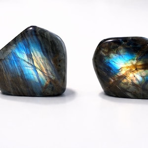 Może przedstawiać: Dwa polerowane kamienie labradorytu z opalizującymi niebieskimi, złotymi i szarymi odcieniami. Kamienie mają gładkie, zaokrąglone kształty i są prezentowane na białym tle. Naturalne przedmioty dekoracyjne.
