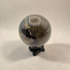 Puede incluir: Una esfera de piedra pulida con patrones en espiral de colores gris oscuro, marrón y crema con fisuras naturales visibles. La esfera se apoya sobre un pequeño soporte negro ornamentado con tres pies decorativos y detalles intrincados.