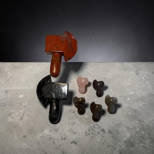 May include: A collection of carved stone axe figurines in various sizes and colors. The largest axe is reddish-brown, while the other axes are black, pink, and brown. The axes are displayed on a gray surface against a dark background.