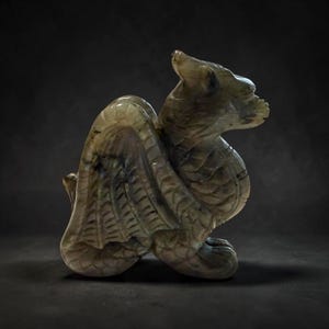 以下が含まれることがあります： 座った姿勢のドラゴンの詳細な彫刻。 ドラゴンは、グレー、グリーン、紫の色合いの石から彫られています。 翼が広げられ、体は複雑に詳細に描かれています。