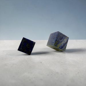 以下が含まれることがあります： 深い青色のラピスラズリの石から彫り出された2つの立方体。金色の黄鉄鉱のインクルージョンが見られます。小さい方の立方体は左側に、大きい方の立方体は右側に配置されており、どちらも天然の鉱物模様が特徴です。