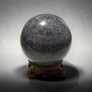 May include: A polished, speckled sphere in shades of purple, green, and gray, resting on a carved wooden stand. The sphere has a reflective surface, and the stand is a dark brown color.