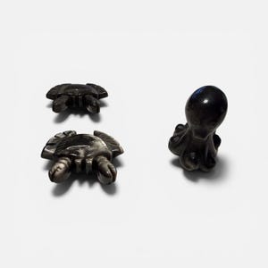 May include: Three dark gray stone figurines: two crabs and one octopus. The crab figurines are positioned at the bottom and top left, while the octopus is on the right. The figurines are on a white background.
