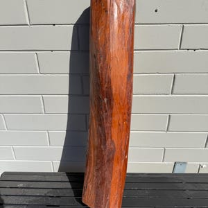 May include: A tall, roughly hewn wooden pillar with a rich, reddish-brown hue. The pillar has a natural, textured surface and a rectangular shape. It stands upright against a light grey brick wall, suggesting a rustic or natural aesthetic.