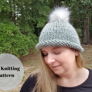 May include: A knitted beanie in shades of gray-green with a fluffy white pom-pom. The hat has a folded brim and is made with a thick yarn. The image includes the text "PDF Knitting Pattern".