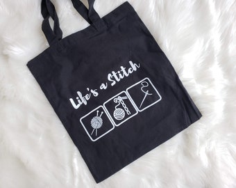 May include: Black tote bag with white text and graphics. The text reads "Life's a Stitch" above three square icons depicting yarn, knitting needles, and a needle and thread. The bag has two black handles.