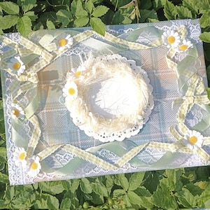 May include: A decorative craft piece with a central nest-like structure on a doily, framed by ribbons, lace, and daisy accents. The ribbons are a mix of solid green and a light green gingham pattern. The background has a plaid design.