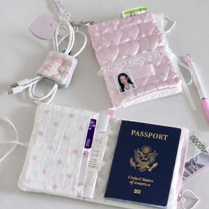 Könnte beinhalten: Ein Reisepasshalter mit einem US-amerikanischen Reisepass, einer rosa gesteppten Reisepasshülle mit einem Foto und einer weißen Stoffhülle mit rosa Sternen. Ein USB-Kabel ist in einem rosa-blauen Stoff mit einem Stern verpackt.