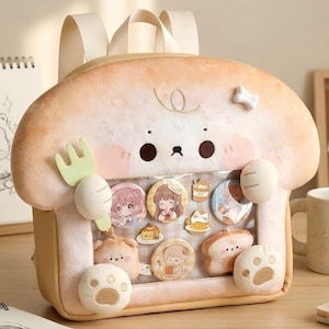 May include: A novelty backpack in the shape of a bread loaf, with a cartoon dog design and decorative badges. The backpack is light beige with white details, a handle, and adjustable straps. The badges inside the clear window feature cartoon illustrations.