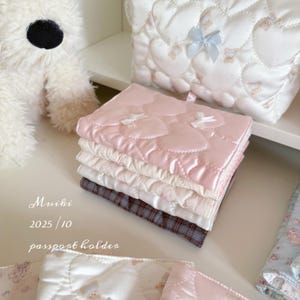 May include: A stack of passport holders in various colors, including pink, white, and a plaid pattern. The pink holder features a heart-shaped quilted design with small white bows. The text "Muiki 2025/10 passport holder" is visible.