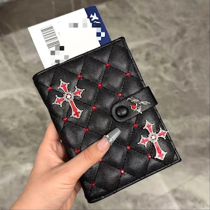 May include: Black quilted passport holder with red cross and stud embellishments. Features a bird clasp and a ticket peeking out. The passport holder is approximately 5 inches tall and 4 inches wide.