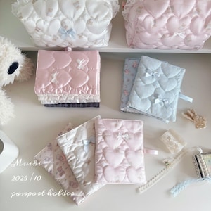 May include: Various passport holders and small bags in pastel shades. The passport holders feature a quilted heart design and small bows. The bags and passport holders are in white, pink, and blue. The text "passport holders" is visible.