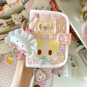 May include: A pink and white gingham patterned notebook cover with a cartoon cat face and the Japanese characters for "happiness". A white plushie with a pink bow is attached. Decorative musical notes and stars are also present.
