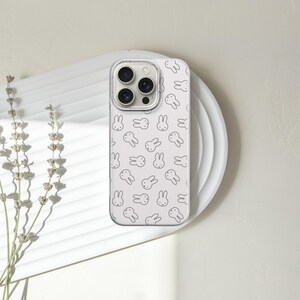 Puede incluir: Una funda de tel&eacute;fono blanca con un patr&oacute;n repetido de caras de conejos negros. La funda del tel&eacute;fono est&aacute; sobre un plato blanco, con una ramita de lavanda seca a la izquierda. El fondo es de color blanco roto neutro.