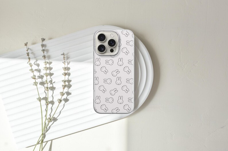 Puede incluir: Una funda de tel&eacute;fono blanca con un patr&oacute;n repetido de caras de conejos negros. La funda del tel&eacute;fono est&aacute; sobre un plato blanco, con una ramita de lavanda seca a la izquierda. El fondo es de color blanco roto neutro.