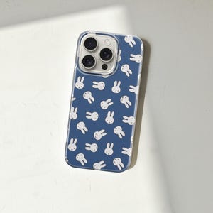 Puede incluir: Funda transparente para tel&eacute;fono con fondo azul y un patr&oacute;n de caras de conejos blancos. La funda tiene un borde transparente y cubre la parte trasera y los laterales del tel&eacute;fono, con recortes para la c&aacute;mara y los botones.
