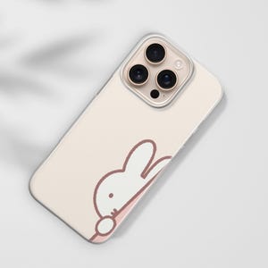 Puede incluir: Funda de tel&eacute;fono rosa claro con un dise&ntilde;o de conejo de dibujos animados. La funda tiene un borde transparente y un recorte para tres lentes de c&aacute;mara. El conejo es blanco con un contorno marr&oacute;n y un acento rosa.