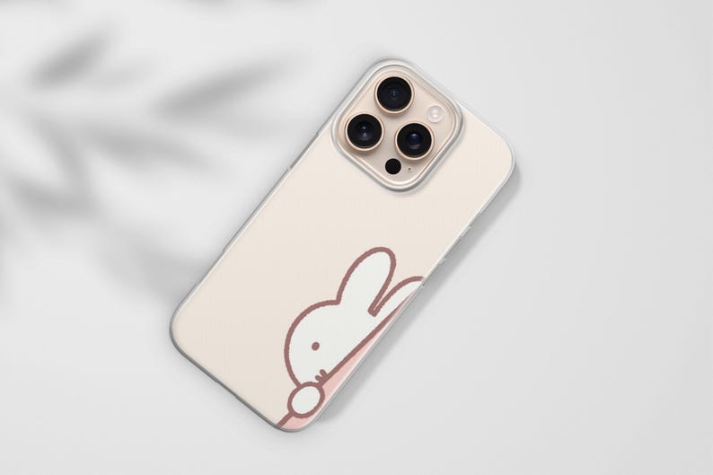 Puede incluir: Funda de tel&eacute;fono rosa claro con un dise&ntilde;o de conejo de dibujos animados. La funda tiene un borde transparente y un recorte para tres lentes de c&aacute;mara. El conejo es blanco con un contorno marr&oacute;n y un acento rosa.