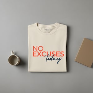 Puede incluir: Camiseta color crema con las palabras "NO EXCUSES Today" en rojo y azul marino. Una pequeña taza de cerámica y un libro cerrado están junto a la camiseta sobre una superficie gris. La camiseta está doblada.