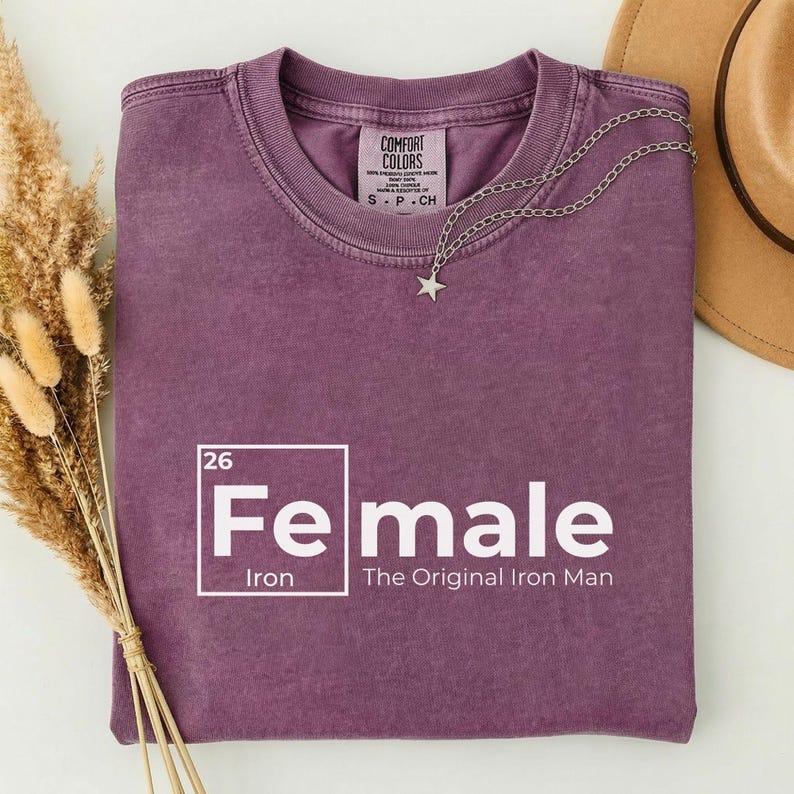 May include: A purple t-shirt with the text "Fe male" in a white, periodic table-style design. The shirt also features the text "The Original Iron Man". A silver necklace with a star pendant rests on the shirt.