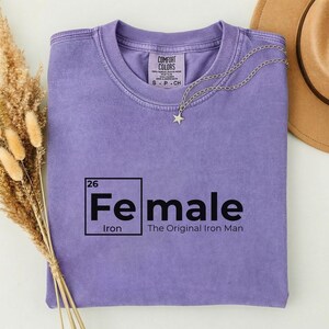 May include: A lilac-colored t-shirt with the text "Female" in a black, periodic table-style font, with "Iron" below. The phrase "The Original Iron Man" is printed below the word "Female". A silver necklace with a star pendant is draped on the shirt.