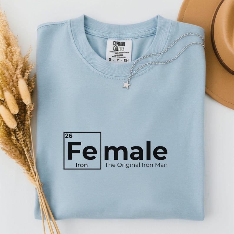 May include: A light blue t-shirt with the word "Female" in a black, periodic table-style font. The text "The Original Iron Man" is below the word "Female". A silver necklace with a star pendant and a brown hat are also visible.