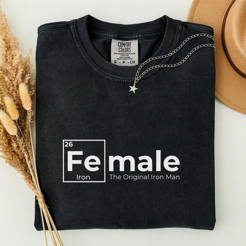 May include: Black t-shirt with the word "Female" in a white, square-framed design, with "Iron" below and "The Original Iron Man" underneath. A silver star necklace is draped across the shirt. The shirt is folded, with a brown hat visible in the background.