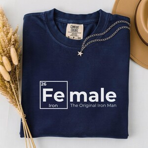 May include: Navy blue t-shirt with the word "Female" in a white, periodic table-style design, with "Iron" below and "The Original Iron Man" beneath. A silver star necklace and a tan hat are visible.