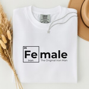 May include: A white t-shirt with the word "Female" in a black, periodic table-style design. The text "Iron" is below the word "Female", and "The Original Iron Man" is below that. A silver star necklace is draped on the shirt.