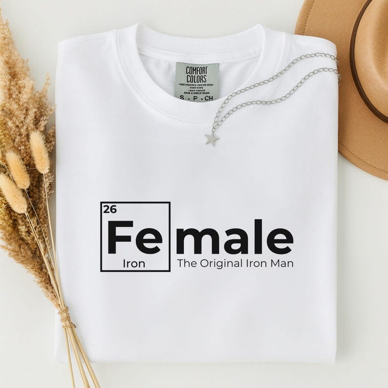 May include: A white t-shirt with the word "Female" in a black, periodic table-style design. The text "Iron" is below the word "Female", and "The Original Iron Man" is below that. A silver star necklace is draped on the shirt.