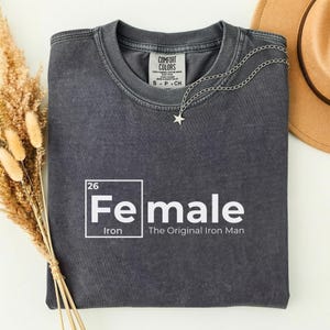 May include: A dark gray t-shirt with the word "Female" in white, styled like a periodic table element, with "Iron" below. A silver star necklace and a brown hat are also visible.
