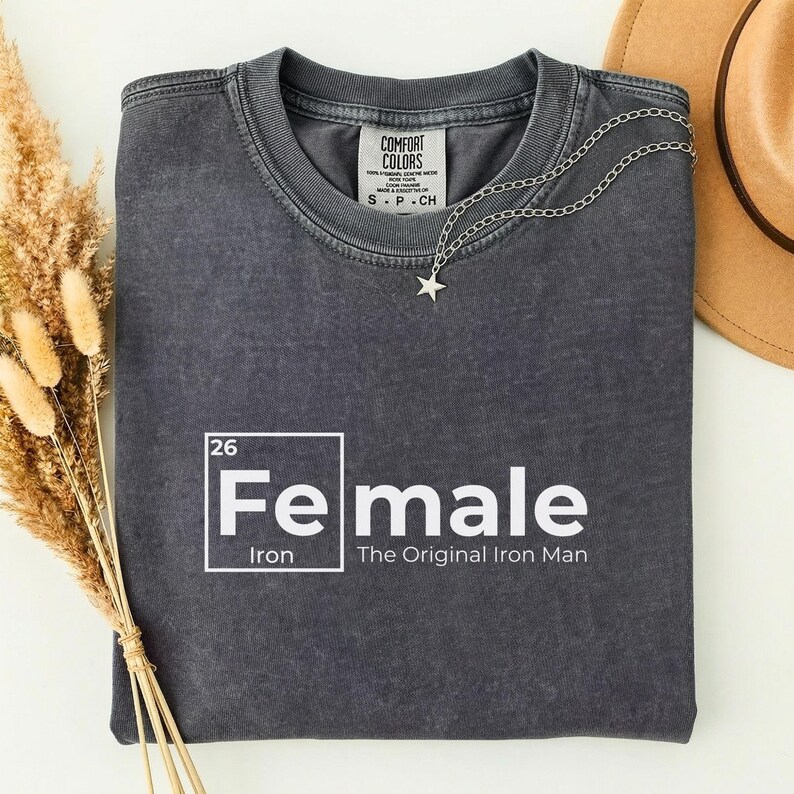 May include: A dark gray t-shirt with the word "Female" in white, styled like a periodic table element, with "Iron" below. A silver star necklace and a brown hat are also visible.