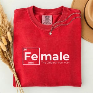 May include: A red t-shirt with the word "Female" in white, with the chemical symbol for iron. The text "The Original Iron Man" is below the word "Female". A silver necklace with a star pendant is draped on the shirt.
