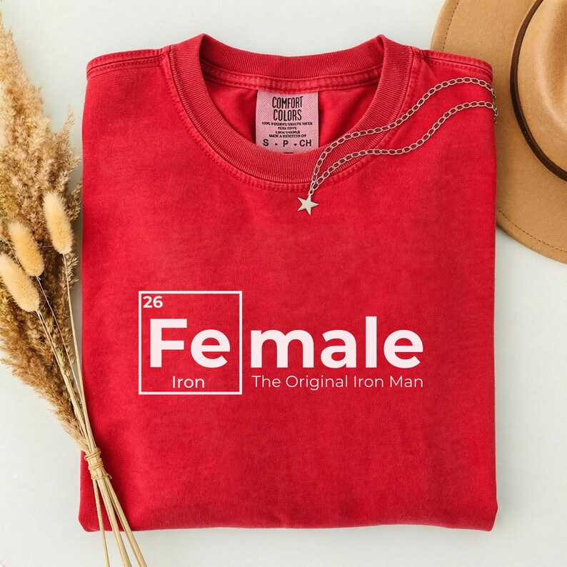 May include: A red t-shirt with the word "Female" in white, with the chemical symbol for iron. The text "The Original Iron Man" is below the word "Female". A silver necklace with a star pendant is draped on the shirt.