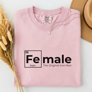 May include: A light pink t-shirt with the word "Female" in a periodic table style design. The text "The Original Iron Man" is below the word "Female". A silver necklace with a star pendant is on the shirt.
