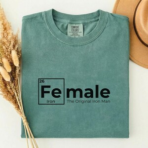 May include: A teal-colored t-shirt with the text "Fe male" in a black, periodic table-style design. The text "Iron" and "The Original Iron Man" are also printed on the shirt. A tan hat is visible in the background.