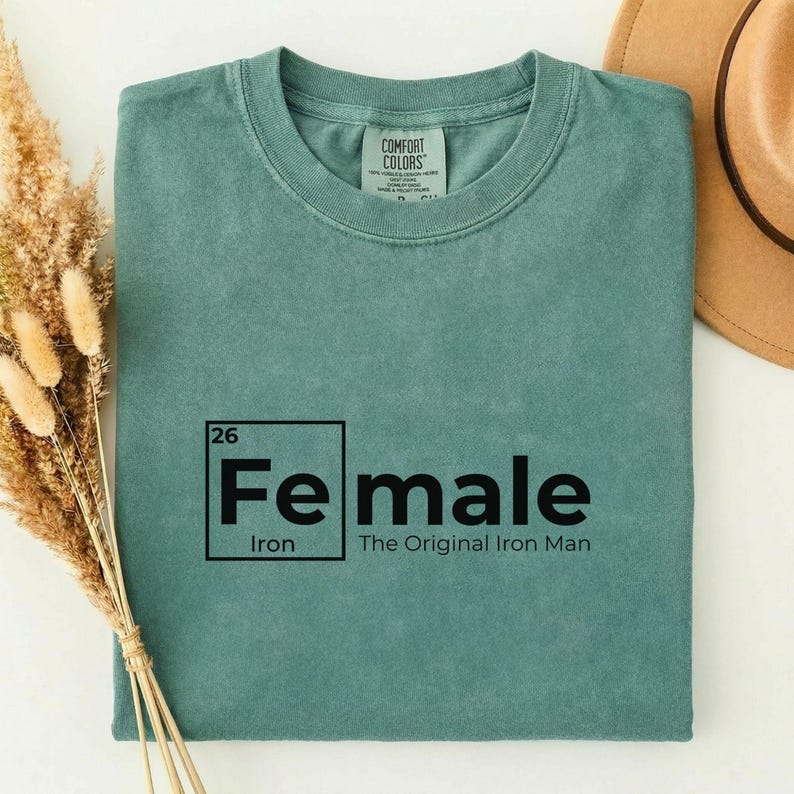 May include: A teal-colored t-shirt with the text "Fe male" in a black, periodic table-style design. The text "Iron" and "The Original Iron Man" are also printed on the shirt. A tan hat is visible in the background.