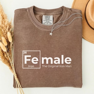 May include: A brown Comfort Colors t-shirt with the word "Female" in white, styled like a periodic table element, with the text "The Original Iron Man." A silver star necklace and a tan hat are also visible.