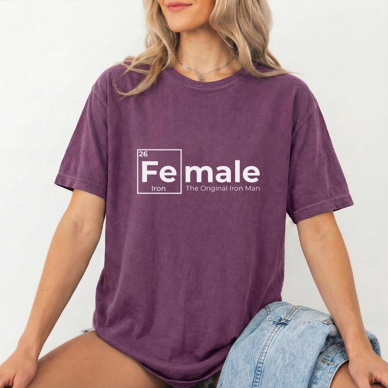 May include: A purple t-shirt with the text "Female" in a white, periodic table-style design. The text "Iron" and "The Original Iron Man" are below the word "Female". The shirt is a solid color and has short sleeves.