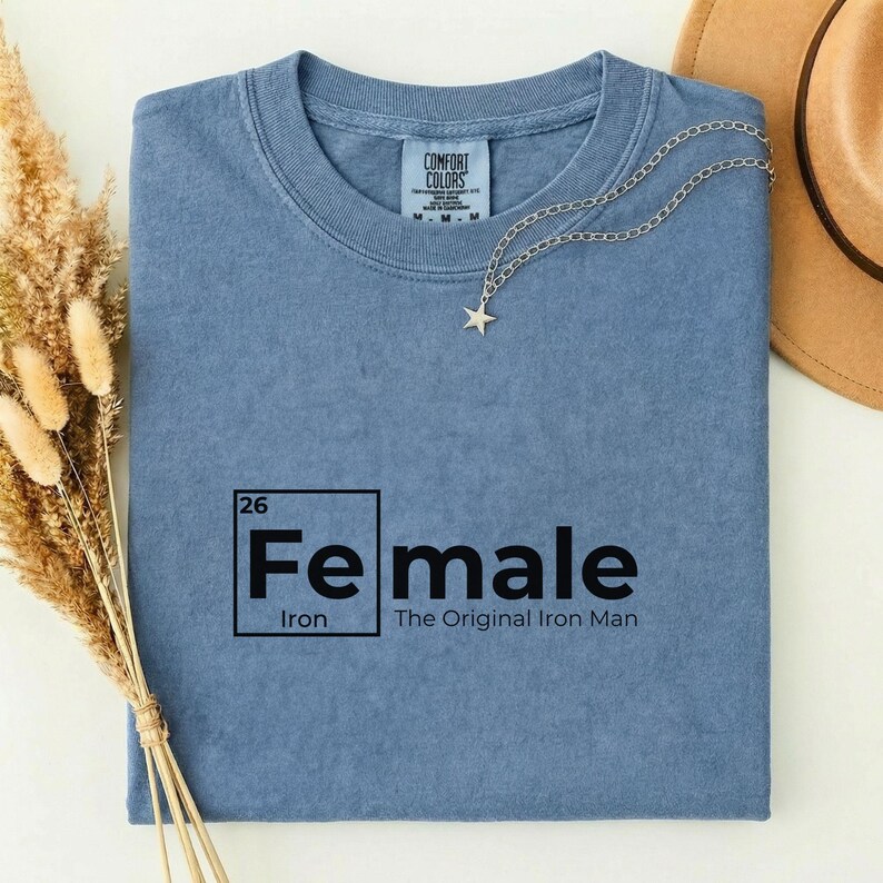 May include: A blue t-shirt with the word "Female" in a black box, with the word "Iron" below it. The text "The Original Iron Man" is below the word "Female". A silver necklace with a star pendant is on the shirt.