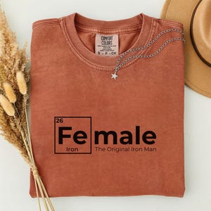 May include: A rust-colored t-shirt with the word "Female" in a periodic table style, with "Iron" below and "The Original Iron Man" beneath. A silver star pendant necklace is draped on the shirt.