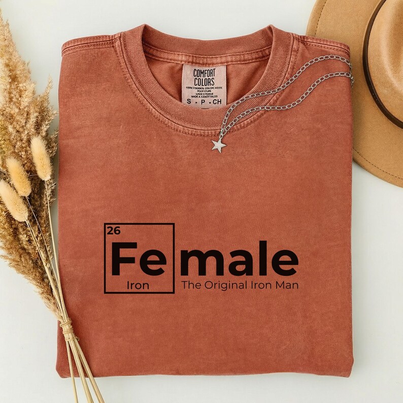 May include: A rust-colored t-shirt with the word "Female" in a periodic table style, with "Iron" below and "The Original Iron Man" beneath. A silver star pendant necklace is draped on the shirt.