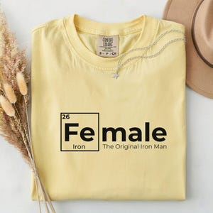May include: A light yellow t-shirt with the word "Female" in a periodic table style, with "Iron" below and "The Original Iron Man" beneath. A silver star necklace and a hat are on the shirt.